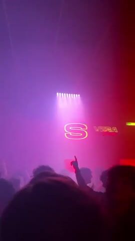 The video shows a crowded indoor scene, likely a nightclub, with attendees dancing in the foreground.  The lighting is primarily red and pink, with a bright neon sign that features the letter "S" and the word "VERA."  The sign is elevated above the crowd.  There is haze or smoke in the air, and moving lights scan across the room.  The focus is on the visual atmosphere and the overall sense of a lively party.  The attendees are dark silhouettes, suggesting they are facing the stage or the source of the music.