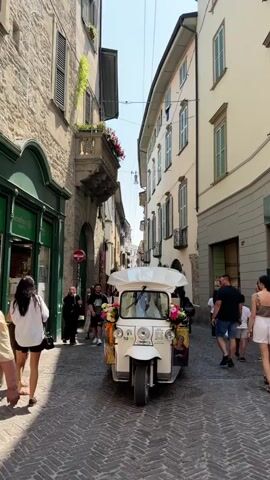 The video shows a narrow cobblestone street lined with old buildings. A white tuk-tuk decorated with flowers is driving down the street. People are walking on the sidewalks and some are looking at the tuk-tuk. The woman in the tuk-tuk is wearing a light pink dress and has blonde hair. The man driving the tuk-tuk is wearing a white shirt. The weather is sunny.