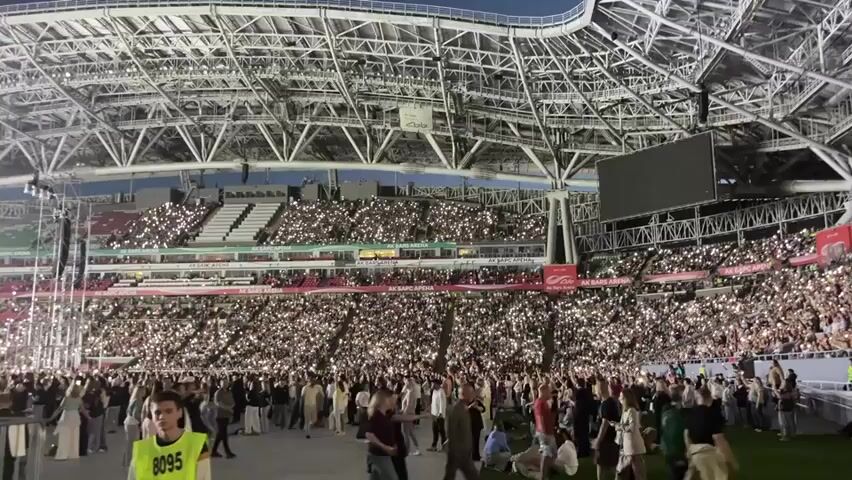 The video captures a wide shot of a large, illuminated stadium filled with a dense crowd of people. The spectators are holding up their mobile phone flashlights, creating a sea of white lights that illuminates the stands. The stadium has a modern, arched roof structure. In the foreground, several individuals are visible, some walking and others standing. One person in the lower left corner is wearing a bright yellow vest with the number 8095 printed on it, indicating they may be security personnel. The overall atmosphere appears to be that of a concert or a large event, given the coordinated use of lights by the audience. The lighting suggests it is evening or night, as the sky above the stadium roof is dark.