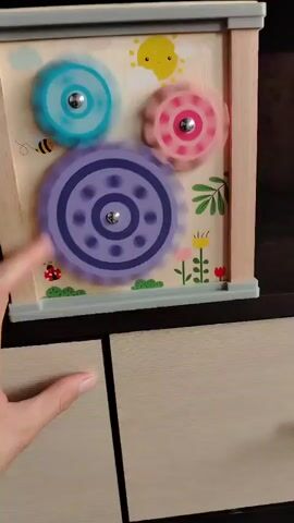 The video shows a wooden toy with three colorful gears. A hand enters the frame and touches the purple gear, causing it to spin. The blue and pink gears also begin to spin in unison. The background of the toy features a drawing of a sun, a bee, plants, and flowers. The toy is positioned on top of what appears to be a wooden cabinet.