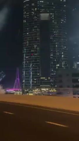 The video shows a night scene, likely in Dubai, as seen from a moving car on a highway.  A tall, unique skyscraper with a large gap in its upper half dominates the view, illuminated by numerous lights from its windows. The building has a block-like, rectangular design.  In the background, a Ferris wheel is visible, lit up in pink and purple hues. A sign that reads "EMAAR" is prominently displayed along the base of the buildings. The foreground shows a concrete barrier lining the highway, with the road surface dimly illuminated by streetlights. The video is taken from inside a vehicle, capturing some internal reflections on the windshield.