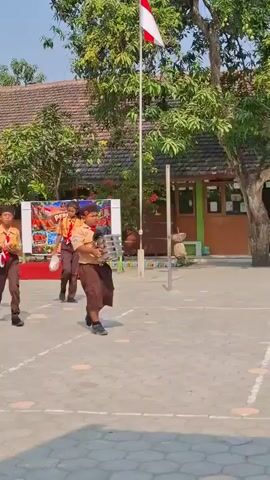 The video shows a group of children in scout uniforms walking in a courtyard. They are carrying various items, including bags and what appears to be musical instruments. There is a flag pole with a flag at the top in the background. The children are walking towards the viewer.