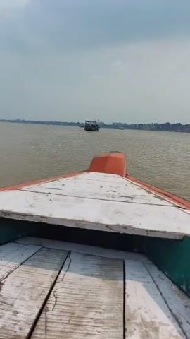 The video shows a first-person view from the front of a boat moving through a wide, murky body of water under an overcast sky. In the distance, there is a city skyline visible on the horizon. Another, larger boat with people on board is seen in front of the camera boat, also moving through the water. The camera boat's wooden deck and orange-tipped bow are visible in the foreground.