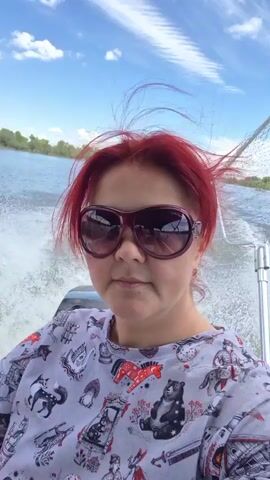 A woman with red hair and sunglasses is filming herself from a boat on a river. She is wearing a t-shirt with various animal and fantasy prints. The boat is moving, creating a wake. The riverbanks are lined with trees. The sky is blue with white clouds. A fishing net is visible in the background.