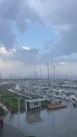 A person in a red shirt and dark pants is walking on a wet paved area near a marina. The marina is filled with many sailboats and yachts. The sky is cloudy with patches of blue. There are also trees and outdoor seating areas visible.