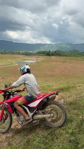 A person wearing a white helmet and a light-colored sweatshirt rides a red dirt bike across a dry, grassy, and dusty landscape. The rider is facing away from the camera and appears to be moving uphill. The terrain is characterized by patches of green grass and bare, reddish-brown earth, with visible tire tracks. In the background, a body of water, possibly a lake or reservoir, is visible, surrounded by dry, cracked earth. Further in the distance, rolling hills and mountains are present, with a cloudy, dramatic sky overhead. The overall impression is one of a rugged, open, and somewhat desolate natural environment.
