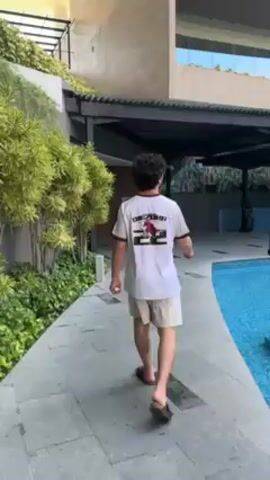 The video shows a man walking along a stone path beside a swimming pool. He is wearing a white jersey with the number 22 on the back, khaki shorts, and sandals. The pool is turquoise blue and surrounded by a black metal structure with a roof. A decorative black birdcage is placed in the pool. The background features a tall building with reflective windows and lush greenery. The camera pans slightly to follow the man's movement and to show more of the surrounding environment. The overall setting appears to be a luxurious resort or hotel.
