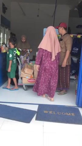 The video captures a moment inside a shop. Four individuals are present. A young boy wearing a green t-shirt and shorts stands on the left, facing away from the camera, with his left leg forward. To his right, a woman in a hijab and patterned clothing holds a piece of paper, looking downwards. In the center, a person with dark hair is partially visible. On the right, a woman in a pink hijab and a long floral dress stands facing away, while a man in a red cap and brown shirt stands next to her. A handcart laden with boxes, some with the "Indomie" logo, is situated in the center of the scene. The environment appears to be a retail space with tiled flooring and shelving visible in the background. A blue mat with the word "WELCOME" is placed on the floor in the foreground. The lighting suggests an indoor environment with natural light entering from an opening.