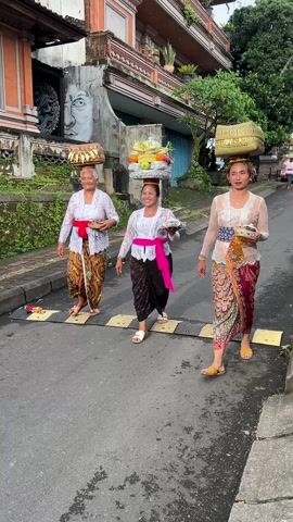 Three Balinese women are walking along a street. They are carrying baskets of offerings on their heads and plates in their hands. The women are wearing traditional clothing, including lace tops, sarongs, and sashes. They are all smiling and looking at the camera. The street is lined with buildings, some of which have traditional Balinese architecture. There are speed bumps on the road. The sky is overcast.