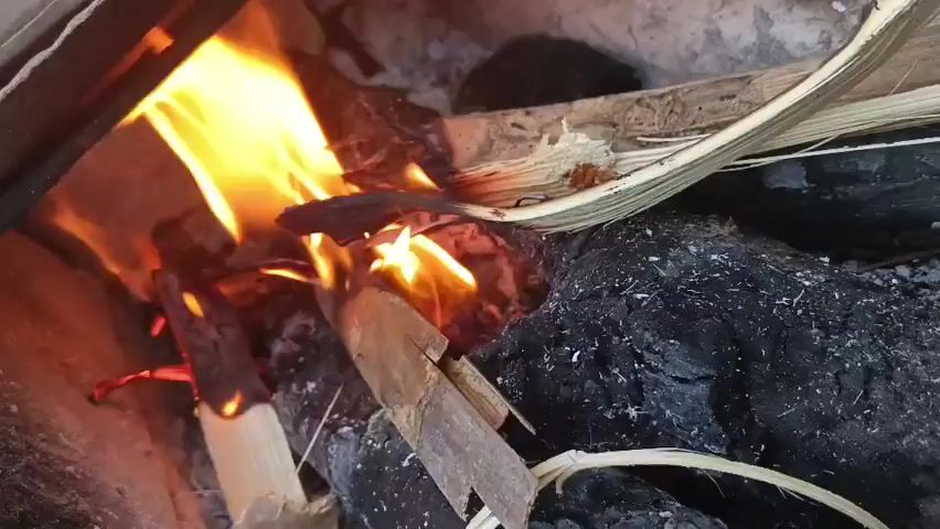 A close-up, still shot shows a small campfire with bright orange flames and white smoke consuming dry, splintered wood and some charcoal. The background is out of focus, but appears to be a rough, earthen surface.