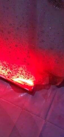 A close-up shot of the inside of what appears to be a washing machine, where clothes are being washed. The water in the machine is a reddish-purple color due to the reflection of a red light. The clothes are not clearly visible, but there is some foam, and glitter is seen swirling around in the water. The camera moves up and down slightly, showing different parts of the water and the glitter. There is a continuous whirring sound of the washing machine throughout the video, and a voice speaking in the background, though what is being said is unclear.
