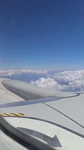 The video shows a view from an airplane window. The wing of the airplane is visible in the foreground, with a section of the engine nacelle. The sky is a clear blue, with a layer of white cumulus clouds below. The camera appears to be panning slightly to the right.