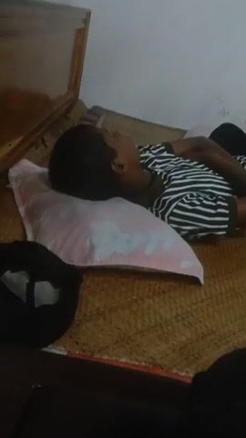 A person lies on their side on a mattress covered with a woven mat. The person is wearing a black and white striped t-shirt and dark pants. A pink pillow with a floral pattern is under their head. The room has white walls and a wooden frame is visible at the top left. In the background, a mirror reflects part of the room, showing a purple curtain and a wooden dresser with a mirror. The lighting is dim.