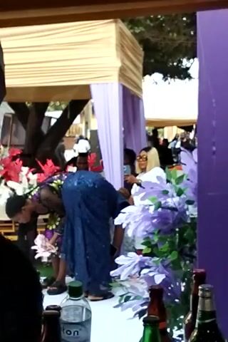 The video depicts a scene outdoors, likely at a gathering or event.  

There are several individuals present, including a woman in a colorful floral dress with a black veil, and another person in a blue sequined outfit. Both are standing near a structure draped in purple and white fabric. The structure appears to have a gold-colored covering on one side. 

There are other people further back, behind the draped structure, but they are not clearly visible. 

In the foreground, there are several bottles of various colors and brands. There are also floral decorations in purple, white, and red. 

The lighting appears to be natural daylight.