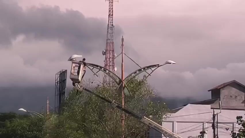 Here's a description of the video:

The video shows a worker in a lift basket performing maintenance on an arched street light fixture. The fixture is made of metal and has several holes along the arches. The lift basket is attached to a crane arm. In the background, there is a tall telecommunications tower, a building with a reddish roof, other street lights, and trees. The sky is overcast with gray clouds. The video focuses on the worker and the light fixture, zooming in slightly.