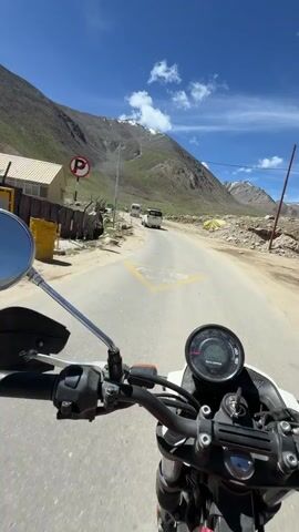 This is a first-person perspective video of someone riding a motorcycle on a paved road in a mountainous, high-altitude region. The rider is wearing a helmet, goggles, and a face mask. The road is winding and appears to be in a sparsely populated area, with some buildings and tents visible on the side of the road. The landscape is characterized by steep, rocky mountains with patches of green vegetation. The sky is blue with some white clouds. There are other vehicles, including a white van and what appears to be a tourist bus, on the road ahead. The rider is focused on the road and the surroundings, with occasional glances at the dashboard of the motorcycle. The video captures the sense of adventure and freedom associated with motorcycle travel in a scenic and remote location.