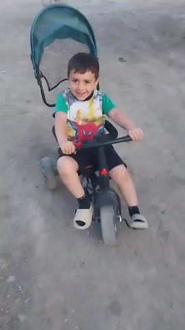 A young boy with short, dark hair and a bright smile is riding a tricycle. He is wearing a green t-shirt with a Spiderman design, black shorts, and white slides with black socks. The tricycle is black with red accents on the handlebars and has a green canopy over the back. The boy is holding onto the handlebars and appears to be pedaling. He is on a dirt path, suggesting an outdoor setting. The lighting suggests it is daytime, possibly late afternoon given the warm tones. The overall mood is cheerful and playful, with the boy looking directly at the camera with an engaging expression. There is no visible text in the image. The scene takes place in Karakert, Armenia.