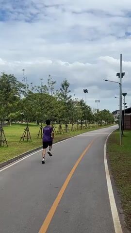 A person wearing a purple shirt and black shorts is running on a paved path with a yellow stripe down the middle. The path is lined with trees and grass. There are lampposts along the right side of the path. The sky is cloudy.