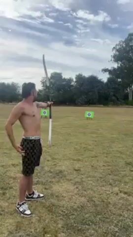 A man is practicing archery at a range. He has a shirtless torso and is wearing black and gold shorts. He is using a recurve bow. After one arrow is shot, the man examines the bow and then passes it to another man. Both men are at the archery range, which has two targets. The setting appears to be outdoors in a grassy field. The sky is partly cloudy.