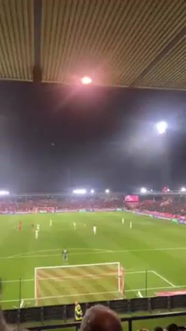 The video shows a soccer game at night. The camera is positioned under the roof of the stadium, looking out over the field. The field is brightly lit with stadium lights. A soccer team in red is visible on the field playing against a team in white. The stands are filled with spectators, mostly wearing red. The goal nets are orange and white.  The camera shakes slightly, indicating it’s handheld. Sounds of the crowd chanting are audible.