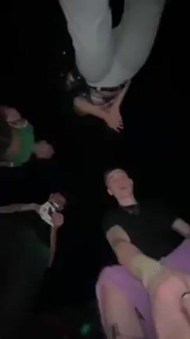 The video shows a group of five people from a low-angle, point-of-view shot, likely taken by someone lying on the ground looking up. The people are gathered close together, forming a circle around the camera. They are all posing and making hand gestures toward the camera. One person is wearing white pants, another a green shirt, and another a lavender sweatshirt. One person appears to be holding a package or item in their hand. The background is dark, suggesting it is nighttime or an indoor location with low lighting. The audio indicates music is playing.