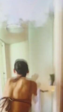 The video shows a woman in a bikini top and pants walking and dancing. She moves through an indoor space with white walls, then through a corridor with vertical white slats. She exits onto a deck with a wooden floor, facing the ocean. The ocean is blue with whitecaps visible. There is a white sunshade above the deck. There is white furniture, including a round couch. The video is somewhat blurry.
