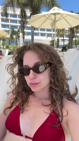 A young woman with curly brown hair and sunglasses is relaxing in a lounge chair outdoors, wearing a red bikini top. She is holding a glass of iced coffee with a straw and drinking from it. In the background, there is a large hotel building, palm trees, white umbrellas, and other people sitting in lounge chairs on a grassy area. The lighting suggests it is daytime, likely sunny.