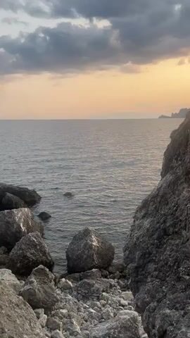 The video shows a rocky coastline with the sea. The camera pans across a cliff face, revealing a small opening in the rocks that leads to a pool of clear water. The sky is overcast with some patches of sunlight breaking through. The sea is calm and the water is a deep blue. There are no people or animals visible in the video.