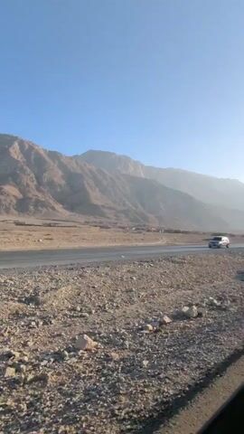 The video shows a landscape with mountains in the background and a flat, rocky terrain in the foreground. A paved road runs through the scene, and a car can be seen driving on it at the beginning of the video. There is also a sign post visible next to the road. The sky is clear and blue. The general atmosphere seems dry and arid. The scene appears to be shot from a moving vehicle.
