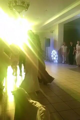 The video shows a dimly lit room with a dance floor. Several people are visible, dancing and interacting. The room is decorated with colored lights that cycle through purple, blue, and green, creating a party atmosphere. There are speakers set up in the room, and a chandelier is hanging from the ceiling. The people are dressed in formal attire, suggesting a special event like a wedding or a formal party. The camera is held by someone who appears to be part of the group, moving with them as they dance.
