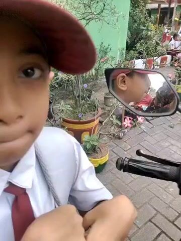 The video begins with a close-up of a child wearing a red and yellow school uniform cap and a white shirt with a red tie. The cap has an emblem and text on it. The child's face is partially visible, and they appear to be looking down. The camera then pans slightly, revealing a motorcycle handlebar in the foreground. In the reflection of the motorcycle's side mirror, another child is visible, also wearing a school uniform. The background shows a school environment with green buildings, potted plants, and a paved walkway. Several other children in school uniforms are seen in the background, some standing and others sitting. One child is riding a motorcycle in the distance. The overall scene suggests a school setting, possibly during a break or dismissal.