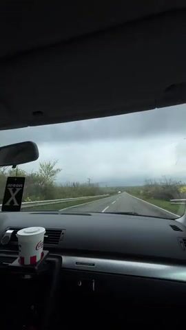 The view from inside a car driving down a rural road is shown. A Coca-Cola cup sits in the car’s center console. A rearview mirror and a hanging air freshener are visible. The car is on an asphalt road in good condition, with green fields and trees on either side. A white van is visible in the distance, driving in the same direction. The sky is overcast.