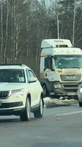 The video shows a white MAN truck that appears to be stuck on the side of a road in the snow. The truck is positioned at an angle, with its front wheels partially submerged in the snow. The road appears to be busy, with cars passing by. The environment is a winter landscape with bare trees in the background. A few other vehicles pass by, including a white car, a grey car, and a yellow taxi. The focus is on the stuck truck and the surrounding traffic.