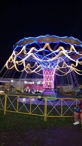 At night, a brightly lit carnival swing ride spins in a circle.  The ride has a center column decorated with pink and white lights in a latticework pattern and a top piece decorated with blue and white lights in a swirling pattern.   Swing seats hang down from the top, spinning around the center.  Behind the ride, there is another carnival attraction.  A yellow and white fence surrounds the base of the ride.   In the foreground, a person in a red jacket and sneakers sits on a bench with a small child in a red outfit.  The area around the bench is grassy.