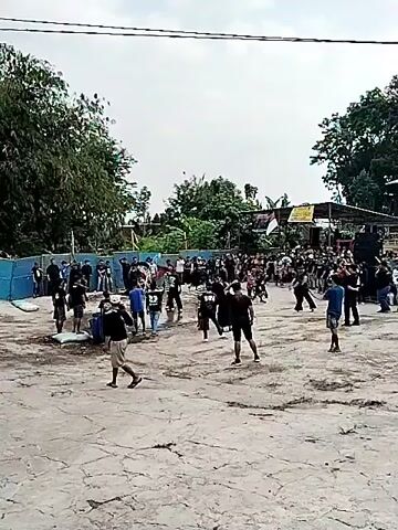 The video shows an outdoor gathering of a large group of people, predominantly young males, in a dusty, open area. Many are dressed in black clothing, some with distinctive designs or logos. The atmosphere appears somewhat chaotic, with individuals moving around and some appearing to be engaged in performances or displays. In the background, there is a structure with a roof, possibly a stage or shelter, adorned with an Indonesian flag and a yellow banner. Trees and foliage surround the area, suggesting a rural or semi-urban setting. Several large blue barrels are visible to the left, and some of the individuals are gathered near them. The overall impression is of a cultural or community event, possibly involving some form of martial arts or dance performance, given the way some individuals are positioned and the energy of the crowd.