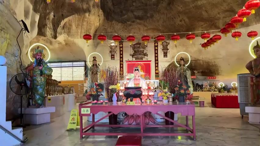 This video shows the interior of a cave temple. In the center, there is a red altar with a large black incense burner and several small candles. Behind the altar, there are statues of deities. To the left and right of the central altar are larger statues of deities. Red lanterns are strung across the ceiling of the cave. The cave walls are visible and appear to be rough and natural. There are also some modern elements such as a fan on the left and a cooling unit on the right. The floor is tiled.