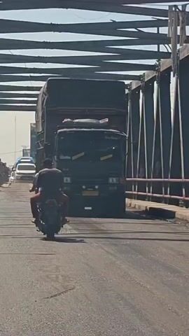 A dark colored truck is seen driving on a bridge. The truck is covered with a dark tarp. In front of the truck, a person is riding a motorcycle. The motorcycle is dark colored and the person is wearing a dark colored shirt and helmet. There is a bridge railing on the right side of the video. The sky is clear and blue. The road surface is asphalt and appears to be wet in places. The truck has a license plate that is yellow and black. The truck also has some yellow tape on its windshield.