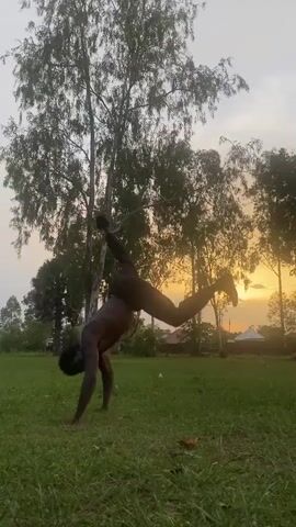 A shirtless Black male is captured mid-air on a grassy field, performing what appears to be an acrobatic flip. The individual is oriented horizontally, parallel to the ground, with one leg extended and the other bent. The background features several trees, some of which are silhouetted against a sky with colors indicating either a sunrise or sunset. In the distance, there are structures that appear to be buildings. The lighting suggests a time near dawn or dusk.
