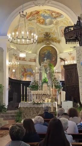The video shows an indoor shot of a church filled with people attending a service. The camera is positioned at the back of the church, facing towards the altar, where a statue of the Virgin Mary is prominently displayed amid flowers and candles. The altar is adorned with detailed decorations and framed by an arch with paintings of religious figures. Above the altar, a large circular painting shows another religious scene. The interior is illuminated by chandeliers, enhancing the ornate details of the church. The churchgoers, a mixed group of adults, are seated in pews. A man in light blue clothing walks along the aisle. The overall atmosphere is serene and reverent.