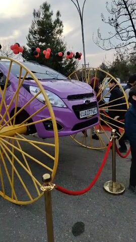 A purple compact car is creatively displayed with yellow wagon wheels.  The wheels are much larger than the car and are positioned in front and behind the vehicle, making it appear as though the car is rolling on the oversized wheels. The car is on display behind a red rope barrier, suggesting it’s part of an event. Above the car, black and red balloons are arranged in small clusters. Two people, including a child, are by the car, seemingly interacting with the display.  The background features a park-like setting with trees and a somewhat cloudy sky. The setting implies a public space, potentially a fair or exhibition.
