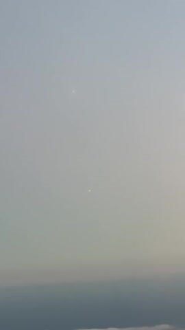 A hazy, overcast sky dominates the video, with a muted palette of grays and pale blues. The sun is visible as a small, reddish-orange disc, appearing somewhat diffused by atmospheric conditions. In the upper portion of the frame, a fainter, whiter spot suggests another celestial body or a brighter patch of haze. The bottom of the frame shows a blurred, indistinct line of what appears to be clouds or the horizon. The video is static, with no discernible movement or action, other than the slight atmospheric diffusion of light.