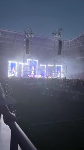 The video shows a wide shot of a large outdoor concert venue at dusk. A stage is set up with scaffolding, bright white lights, and large screens displaying images of performers. Smoke machines are active, creating a hazy atmosphere. A large crowd of people is visible in front of the stage, behind barriers. The sky is a deep blue, transitioning to night. The camera appears to be held by someone in the audience, and their shoulder and hooded head are partially visible in the foreground, suggesting a first-person perspective of the concert. The overall scene depicts a lively and energetic event.