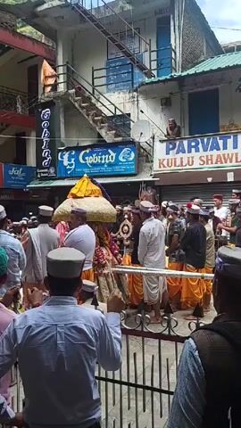 A lively religious procession is captured on a bright, sunny day in what appears to be a street or public square in Kasol, India. Numerous people, predominantly men, are gathered, many wearing traditional Himachali caps – round caps with colorful patterned bands – and some in white kurtas paired with white or orange lower garments, suggesting traditional attire.

At the center of the crowd, a decorated deity or religious object, adorned with a large, elaborate yellow and white umbrella-like canopy and rich fabrics, is being carried by several individuals. The procession is accompanied by traditional music, with rhythmic sounds audible. Spectators line the street, with some observing from behind a metal railing in the foreground, while others are closer to the moving procession.

In the background, multi-story buildings with balconies and external staircases line the street. Several shop signs are visible: "PADHA FAMILY GUEST HOUSE RIVER VIEW ROOMS" on a red banner, and multiple signs for "Gobind" (including "G Gobind" and "CG Gobind"). Another sign on the right reads "PARVATI KULLU SHAWL," and a small "WELCOME" sign is visible near the top of a building. The overall atmosphere suggests a cultural or religious event in a Himalayan town setting.