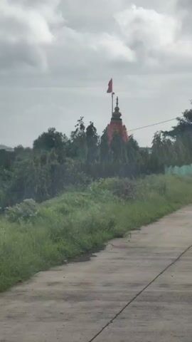 The video captures a glimpse of a temple. The temple has a distinct spire and is adorned with a red flag. It is surrounded by trees, and the foreground shows a dirt road with green vegetation on the side. The sky is overcast with clouds. The camera appears to be moving, suggesting it is filmed from a vehicle.