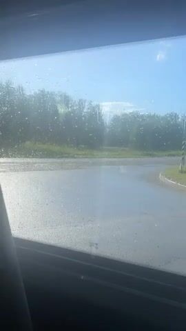 The video shows a view through a window with water droplets on it. Outside the window, there is a wet road with puddles, likely from rain. Trees and green grass are visible in the background under a bright blue sky with some clouds. The sun appears to be shining, creating reflections on the wet surfaces. A black and white striped pole is visible on the right side of the frame. The perspective suggests the video was taken from inside a vehicle.