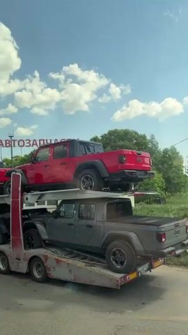 The video shows a trailer carrying multiple Jeeps in various colors. The trailer is loaded with Jeeps stacked on top of each other, and appears to be stopped, with the camera capturing the scene from the side. The cars on the trailer include a red Jeep on the top level, and black and gray Jeeps on the other levels. In the background, there is a sign in what appears to be Russian, reading "AVTOZAPCHASTI" (Автозапчасти), which likely means "auto parts". The sky is blue with some clouds.