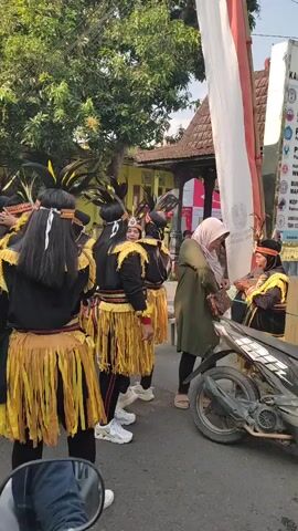 The video shows a group of people in traditional attire, possibly performers, walking outdoors. They are wearing black tops with gold fringe detailing around the shoulders and yellow fringed skirts. Some have headbands with feathers. One person in the foreground is facing away from the camera, adjusting their hair. Another person in the middle ground is looking to the side. Further back, a person in a green hijab and black pants is visible near a parked scooter. The background includes a traditional-looking building with a tiled roof, trees, and a white banner. The scene appears to be a street or a pathway.