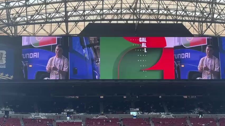 A large video screen in a stadium displays a presentation for Portugal. On the left and right sides of the screen, a man identified as Cristiano Ronaldo is shown standing next to a blue bus with "HYUNDAI" visible on it. In the center of the screen, the word "PORTUGAL" appears in white text with the Portuguese flag on either side. The background of the center section is a gradient of green with red accents. The stadium seating is visible in the foreground, with a large number of spectators in attendance. The stadium has a complex metal roof structure.