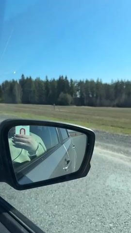 The video, shot from inside a moving car, focuses on the side mirror's reflection and the passing scenery. The mirror shows a person in a green hoodie taking a picture with their phone, the car's door, and a portion of the back seat. Outside the window, a clear, bright blue sky dominates the view, while a field and a line of trees stretch across the horizon. The road is visible in the foreground, showing a gray, coarse surface. The car is moving along a rural route on a sunny day.