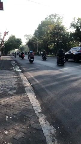 The video shows a street scene with multiple motorcycles and a car. The motorcycles are traveling in the same direction on the road, with some appearing to be in a convoy. The car is also moving in the same direction. The road is paved, and there is a sidewalk on the left side of the frame. Trees line the side of the road, and the sky is bright, suggesting it is daytime. Some of the motorcycles have riders wearing helmets. The overall impression is of traffic on a city street during the day.